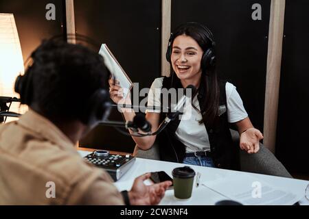 Portait von glücklicher Radiomoderatorin lacht, lauscht männlichen Gästen, Moderatorin und hält ein Drehbuch, während sie eine Live-Show im Studio moderiert Stockfoto