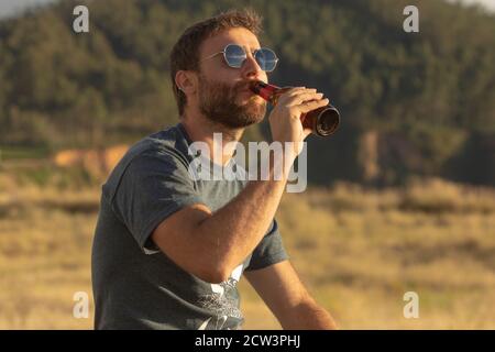 Ein junger Mann, mit Gläsern und Bart, trinkt Bier aus einer Flasche, während er die Landschaft und den Sonnenuntergang in Galicien, Nordspanien, genießt. Stockfoto