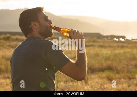 Ein junger Mann, mit Gläsern und Bart, trinkt Bier aus einer Flasche, während er die Landschaft und den Sonnenuntergang in Galicien, Nordspanien, genießt. Stockfoto