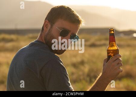 Ein junger Mann, mit Gläsern und Bart, trinkt Bier aus einer Flasche, während er die Landschaft und den Sonnenuntergang in Galicien, Nordspanien, genießt. Stockfoto