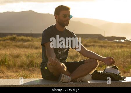 Ein junger Mann, der eine Brille trägt, meditiert in einer Yoga-Haltung, während er die Nachmittagssonne auf einer Promenade in Nordspanien genießt. Stockfoto