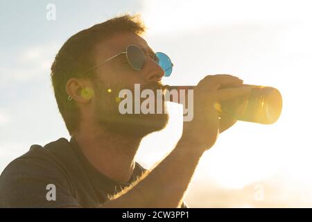 Ein junger Mann, mit Gläsern und Bart, trinkt Bier aus einer Flasche, während er die Landschaft und den Sonnenuntergang in Galicien, Nordspanien, genießt. Stockfoto