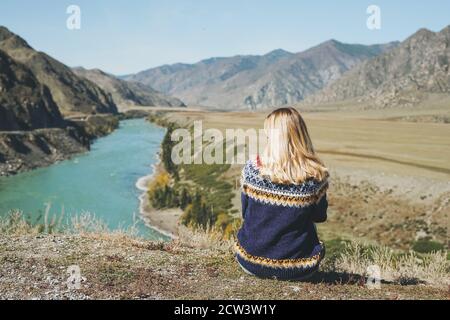 Junge blonde Frau im nordischen Pullover auf dem Hintergrund des türkisfarbenen Katun-Flusses, Altai Berge sitzen Stockfoto