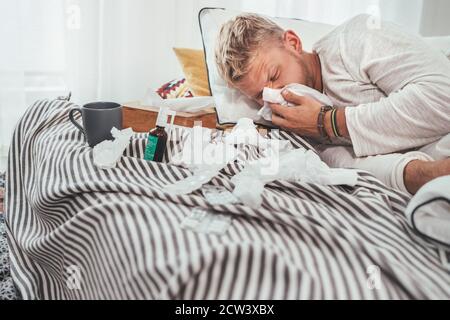 Ungesunder und müder Mann niesen oder wischende notty Nase Mann liegt in einem gemütlichen Bett neben einer Menge von gebrauchten Papiertaschentücher und Medikamente auf der Decke. S Stockfoto