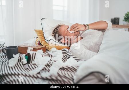 Ungesunder und müder Mann niesen oder wischende notty Nase Mann liegt in einem gemütlichen Bett neben einer Menge von gebrauchten Papiertaschentücher und Medikamente auf der Decke. S Stockfoto