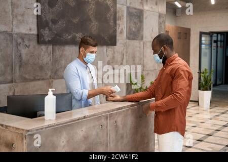 Junge männliche Rezeptionist mit medizinischer Maske an der Rezeption stehend in Die moderne Büro-und Körpertemperatur der afrikanischen Mann messen Mit Infrarot Stockfoto