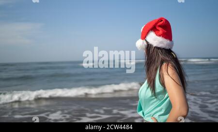 Junge Frau im Weihnachtsmann Hut steht an der Küste bei sonnigem Wetter. Erwachsene Brünette in Santa Claus Hut posiert in der Nähe winkende Meer im Resort Stockfoto