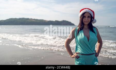 Junge Frau im Weihnachtsmann Hut steht an der Küste bei sonnigem Wetter. Erwachsene Brünette in Santa Claus Hut posiert in der Nähe winkende Meer im Resort Stockfoto