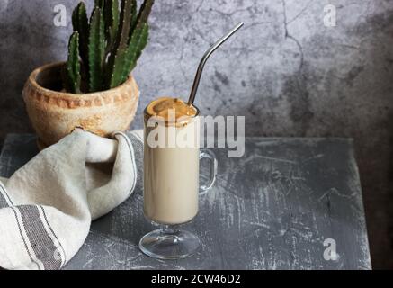 Dalgona Kaffee in einer transparenten Tasse mit einem Cocktail Stroh und Kaktus auf einem Betontisch. Stockfoto