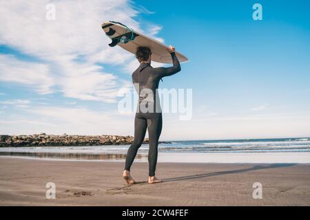 Porträt eines jungen Surfers am Strand, der sein Surfbrett hochhält und einen schwarzen Surfanzug trägt. Sport und Wassersport Konzept. Stockfoto