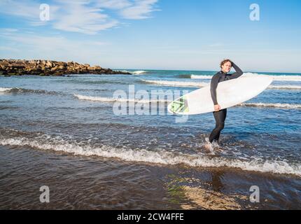 Junger Surfer, der mit seinem Surfbrett in einem schwarzen Surfanzug ins Wasser tritt. Sport- und Wassersportkonzept. Stockfoto