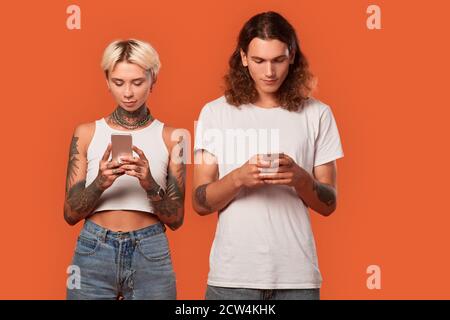 Freistil. Junge tätowierte Paar stehend isoliert auf orange browsing Smartphone konzentriert antisozial Stockfoto