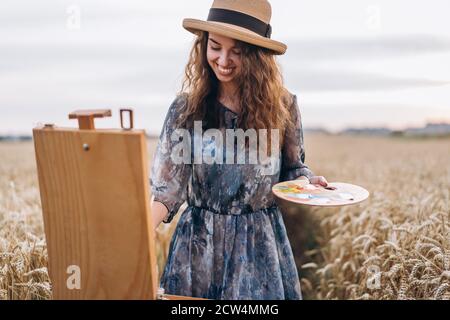 Porträt einer lächelnden Künstlerin mit lockigen Haaren im Hut. Mädchen zeichnet ein Bild von einer Landschaft in einem Weizenfeld. Stockfoto