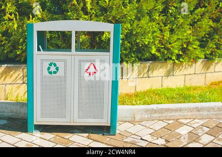 Mülleimer auf der Gasse im Parkbereich. Eisentank für verschiedene Abfallarten. Aufrechterhaltung der Sauberkeit im Erholungsbereich der Menschen. Grüne Tannen hinter einem Ziegelzaun. Stockfoto