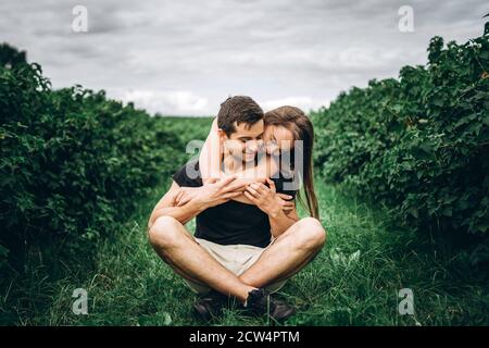 Ein Mann sitzt auf grünem Gras zwischen Johannisbeerbüschen, eine Frau mit langen Haaren umarmt ihn von hinten. Hintergrund der Johannisbeerplantagen. Stockfoto