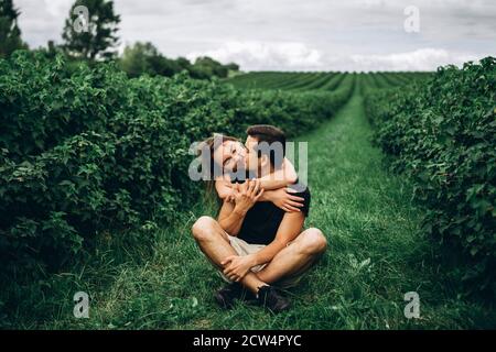 Ein Mann sitzt auf grünem Gras zwischen Johannisbeerbüschen, eine Frau mit langen Haaren umarmt ihn von hinten. Hintergrund der Johannisbeerplantagen. Stockfoto