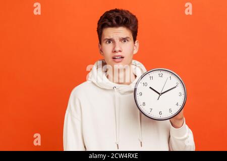 Schockiert Teenager in beiläufigen weißen Hoodie Angst, zu spät, hält in der Hand Wanduhr, Frist, Pünktlichkeit. Innenaufnahme des Studios isoliert auf Orange Stockfoto