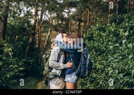 Junges Paar mit Rucksäcken auf dem Rücken im Wald. Liebevoller Mann küsst seine schöne Freundin auf einer Wanderung im Wald. Stockfoto
