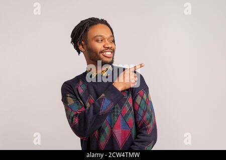 Fröhlicher geheimnisvoller afrikanischer Mann mit Dreadlocks im stylischen Sweatshirt, der mit einem toothy Lächeln auf dem Gesicht den Finger weg zeigt, leerer Raum für Ihre Werbung Stockfoto