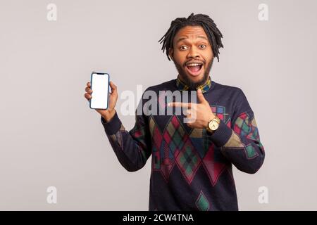 Extrem überrascht afrikanischen Kerl mit Dreadlocks zeigen Finger auf Smartphone leer leer, wunderte sich mit Anwendung erstaunt. Innenaufnahmen im Studio Stockfoto