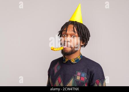 Nahaufnahme afrikanischer Mann mit Dreadlocks trägt Party-Kegelkappe und bläst Party-Pfeife, feiert Jubiläum, Hungout. Innenaufnahme des Studios isoliert auf gr Stockfoto