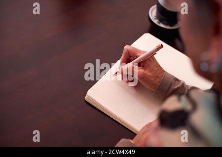 Nahaufnahme Einer Älteren Frau Zu Hause, Die Am Tisch Sitzt Und Schreiben In Notizbuch Oder Journal Stockfoto