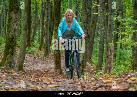 Blonde Mädchen in schwarzen Leggings und einer türkisfarbenen Jacke reitet ein Fahrrad im Herbstwald. Gesunde Lebensweise und aktive Erholung. Stockfoto