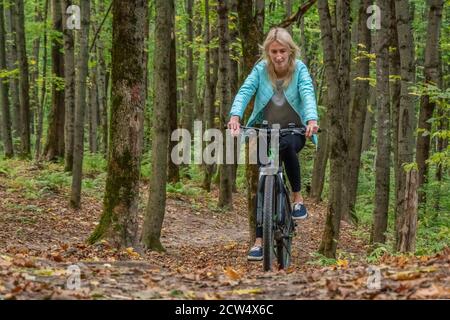 Blonde Mädchen in schwarzen Leggings und einer türkisfarbenen Jacke reitet ein Fahrrad im Herbstwald. Gesunde Lebensweise und aktive Erholung. Stockfoto