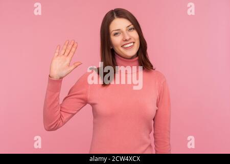 Freundliche positive Frau mit braunen Haaren winkt ihre Hand zeigt hallo Geste, Begrüßung und Begrüßung. Innenaufnahme des Studios isoliert auf rosa Hintergrund Stockfoto