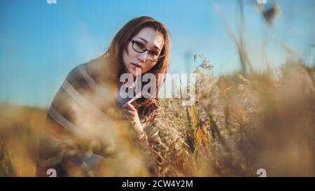 Eine schöne junge mongoloide Frau in einem gestreiften Tuch gekleidet steht inmitten von wildem, hohen Gras auf einem Feld im Sommer, beleuchtet von Sonnenlicht Stockfoto