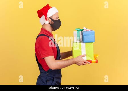Profil Portrait Deliveryman in Uniform, roter weihnachtsmann Hut und chirurgische medizinische Maske mit Paketen und Geschenken, Lieferung in Quarantäne. innenstudi Stockfoto