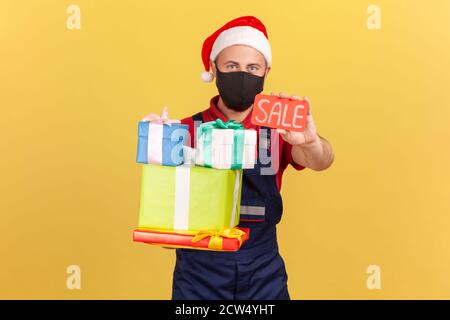 Starke Kurier in medizinischen Schutzmaske, Uniform und weihnachtsmann Hut hält viele Geschenkboxen und zeigt Verkauf Coupon, Weihnachtsverkäufe bei Bestellungen und Deliv Stockfoto