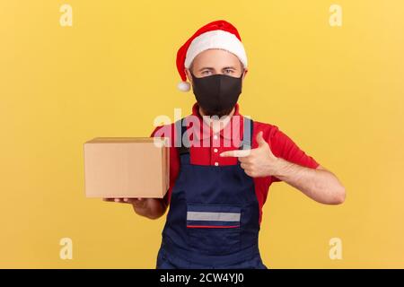 Deliveryman in Uniform und Urlaub weihnachtsmann Hut mit schützenden medizinischen Maske zeigt Finger auf Karton in der Hand, Lieferung in Quarantäne. Stockfoto
