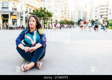 Brunette Frau in legerer Kleidung sitzt auf dem Boden in einem Platz in Europa mit Menschen, Blick in die Ferne. Copy Space Stockfoto