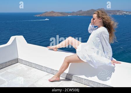 Frau in weißem Kleid Sonnenbrille sitzt auf der weißen Terrasse suchen Meerblick Stockfoto