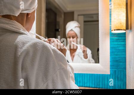 Junge Frau im Bademantel und Handtuch putzen ihre Zähne in Spiegelvorderseite Stockfoto