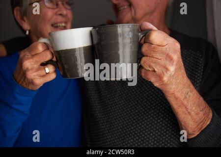 Lächelndes Senior Pärchen, Das Mit Tassen Bei Ihnen Echt Cheers Macht Mit Heißgetränk Zu Hause Stockfoto