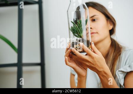 Mädchen, das die Glasflasche mit kleiner Pflanze genau anschaut, hält sie in den Händen Stockfoto