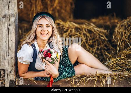 Niedliche Blondine im Dirndl, traditionelles Festkleid mit Blumenstrauß auf dem Holzzaun auf dem Bauernhof mit Heuhaufen Stockfoto