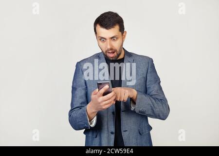 Junger Geschäftsmann Mann mit Bart in einer Jacke mit einem Smartphone erschrocken schockiert mit einem überraschenden Gesicht, erschrocken und aufgeregt mit einem Ausdruck von Angst. Porträt eines Mannes auf grauem Hintergrund. Stockfoto