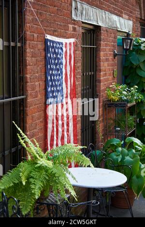 BROOKLYN, NY - SEPTEMBER 23 2020: Eine amerikanische Flagge hängt an der Seite eines alten Backsteinhauses, in einem kleinen Garten zwischen Pflanzen und einem Drahttisch und Stühlen Stockfoto