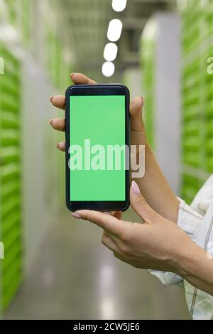 Vertikale Nahaufnahme der weiblichen Hände halten Smartphone mit grün Bildschirm vor dem Hintergrund der Selbstlagereinrichtung Stockfoto