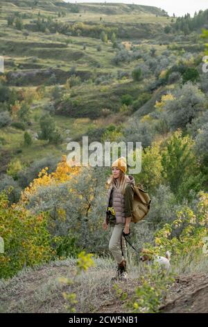 Junge weibliche Backpacker in warmer Casualwear mit ihrem Haustier bewegen Fußweg hinunter Stockfoto