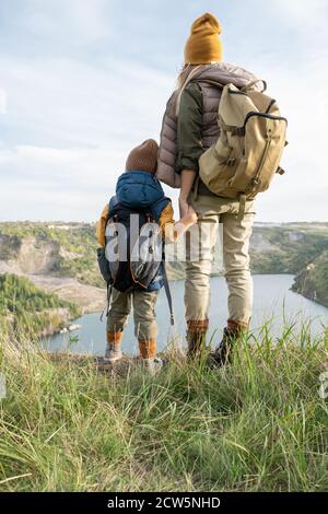 Rückansicht der jungen Mutter und des kleinen Sohnes mit Rucksäcken Am See stehen Stockfoto