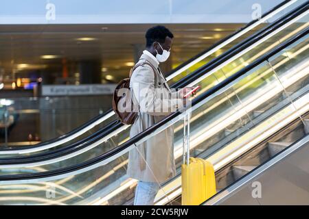 Afro-amerikanischen Reisenden Mann mit gelben Koffer steht auf Rolltreppe in Flughafen-Terminal, tragen Gesicht medizinische Maske, um sich vor Kontakt mit zu schützen Stockfoto