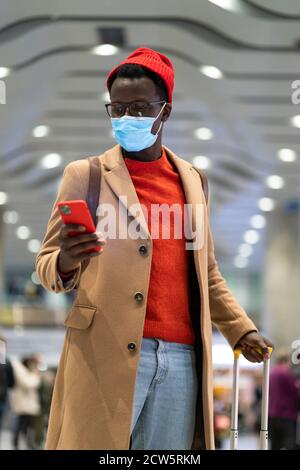 Afroamerikanischer Reisender Mann mit gelben Koffer steht im Flughafen-Terminal, mit Telefon, rufen Taxi, tragen Gesicht medizinische Maske, um sich zu schützen Stockfoto