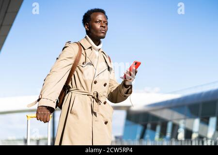 Afro-amerikanische Millennial Traveler Mann mit gelben Koffer steht im Flughafen-Terminal, hält Telefon, anrufen und auf der Suche nach einem Taxi oder Car Sharing. B Stockfoto