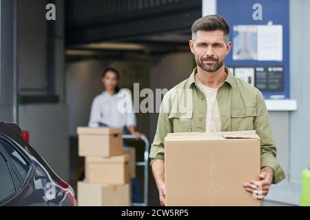 Waist up Porträt von schönen Mann halten Box und Blick auf die Kamera, während Sie bei der Selbstaufbewahren-Anlage stehen, kopieren Raum Stockfoto