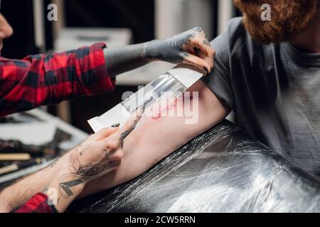 Junge informelle Mädchen Tattoo Künstler setzt einen Verband mit Vaseline Auf der Hand eines jungen Mannes nach erfolgreicher Bewerbung Ein Tattoo auf seiner Hand Stockfoto