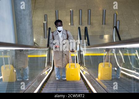 Afro-amerikanischen Reisenden Mann mit gelben Koffer steht auf Rolltreppe in Flughafen-Terminal, tragen Gesicht medizinische Maske, um sich vor Kontakt mit zu schützen Stockfoto
